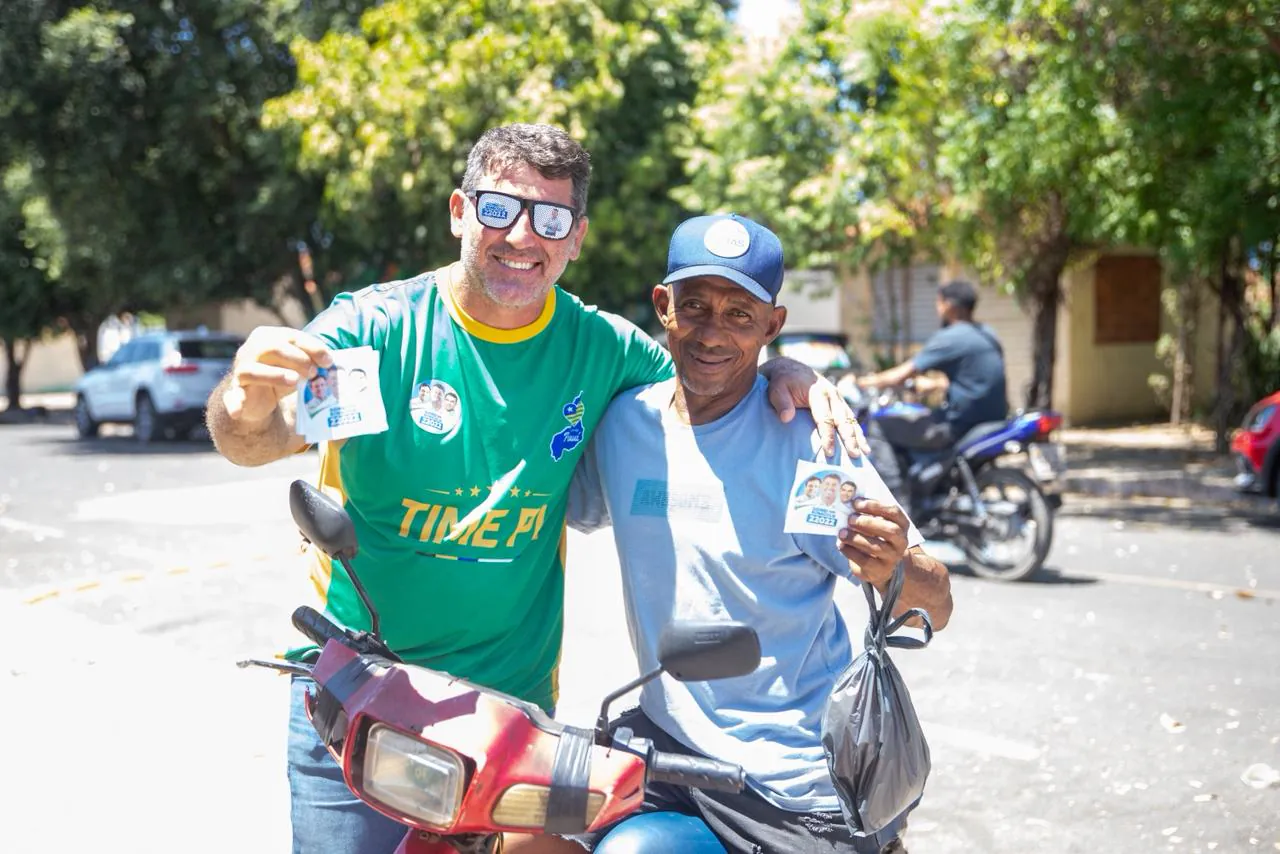 Sidnei Bonacolsi ao lado de um homem sobre uma motocicleta, os dois mostrando santinhos da campanha.